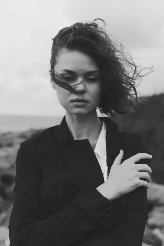 Moody woman in a windblown jacket on a rugged coast, black and white scene. Focused expression, hair and scarf flutter as stormy light adds texture. Artistic, minimalist photography highlighting