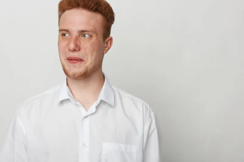 Young man with red hair and freckles wearing white shirt looks sideways with thoughtful expression on plain light background. Concept of casual style, contemplation, and natural appearance.