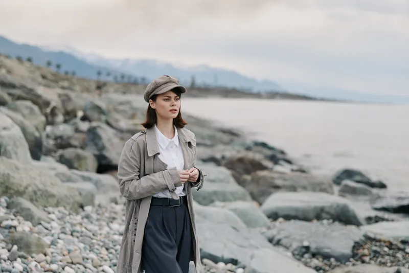 Young woman wearing a patterned coat and hat stands on a rocky beach with mountains and sea in the background, looking thoughtfully into the distance. Overcast sky sets a calm mood.