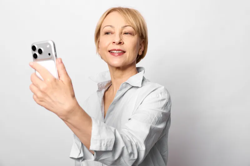 Smiling woman holding smartphone for a selfie on a clean white background, close portrait with relaxed posture, soft studio light, technology and communication concept, daily life
