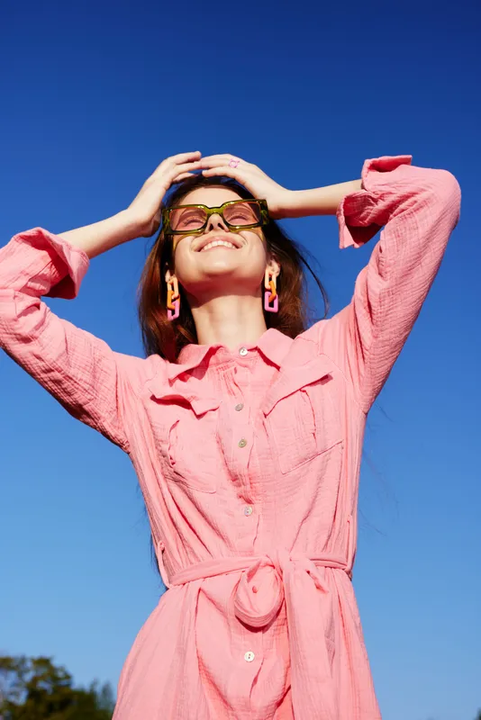 Happy young woman in a pink dress enjoying sunny weather outdoors with a blue sky, showcasing cheerful emotions and vibrant summer fashion