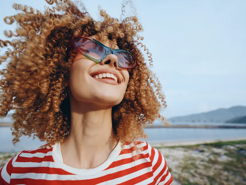 A joyful portrait of a woman with voluminous curly hair, oversized sunglasses, and a bright striped shirt, enjoying a sunny day by the sea.