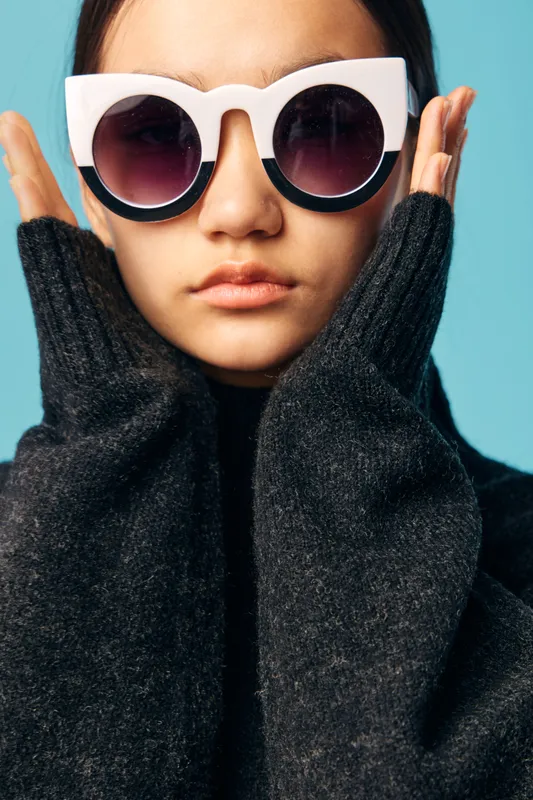 Young woman wearing large white sunglasses and dark knitted sweater posing with hands near face on blue background for fashion, style and portrait photography concept.