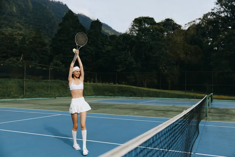 Young female tennis player in a sporty outfit celebrating a victory on a sunny outdoor court, showcasing athleticism and joy while surrounded by lush greenery