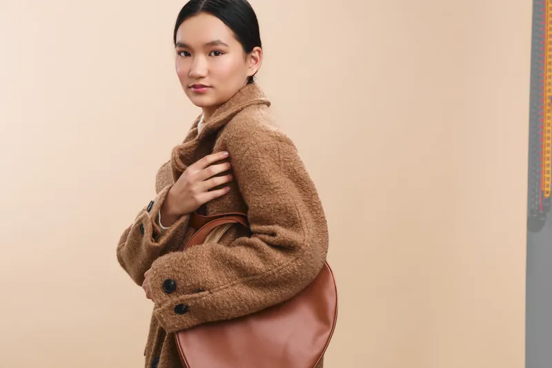 Asian woman in a tan coat holds a brown handbag as she walks in a neutral studio setting, exuding confident urban style and poised elegance with a calm, friendly expression.
