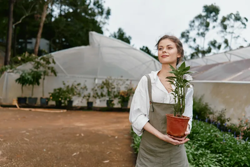 Young woman holding a potted plant in a greenhouse, showcasing a passion for gardening and sustainable living with a serene expression and natural backdrop