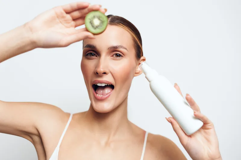 Young woman with blonde hair holds a sliced kiwi fruit near her eye while spraying a white cosmetic bottle on her face in a bright studio setting.