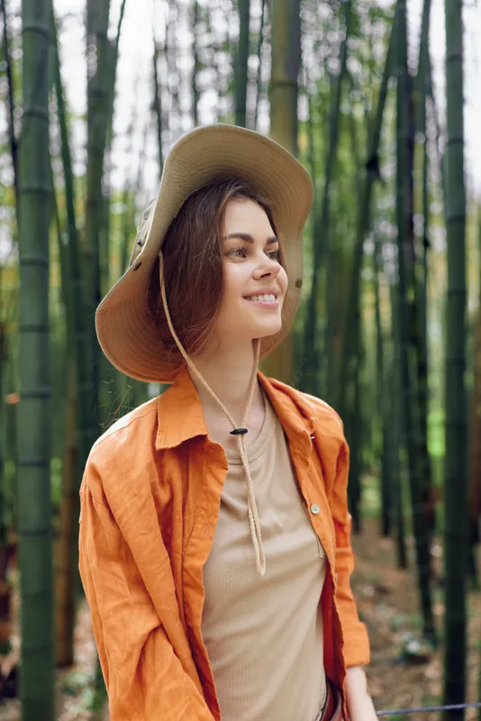 Woman smiling in a bamboo forest wearing a wide brim hat and orange jacket, portrait of nature and travel outdoors with relaxed expression and soft natural light.