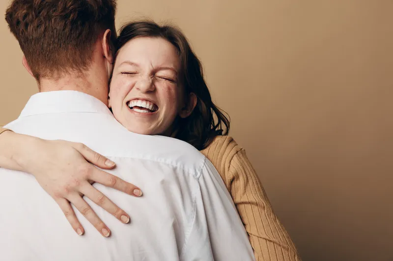 Happy woman with closed eyes embraces man wearing white shirt, smiling joyfully. Warm beige background creates cozy atmosphere. Concept of love, affection, and emotional connection.