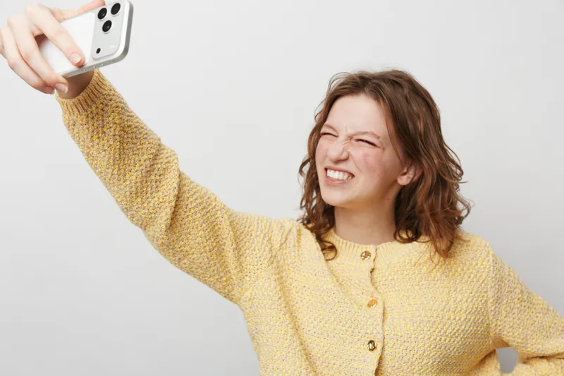 Young woman in yellow sweater takes a selfie with smartphone, making a funny face. Studio shot on white background. Concept of casual lifestyle, technology, and social media.