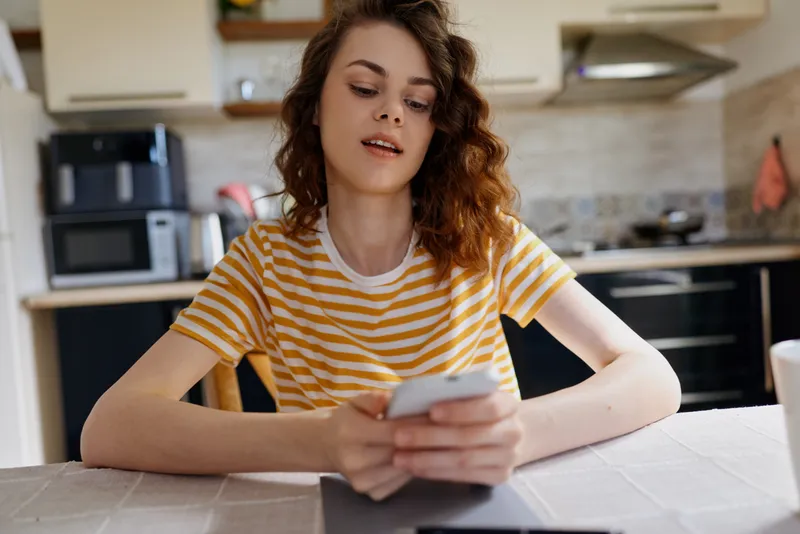 Young woman in a striped t shirt sits at a table in a modern kitchen, looking at a smartphone, relaxed mood, home lifestyle scene, natural light, candid moment