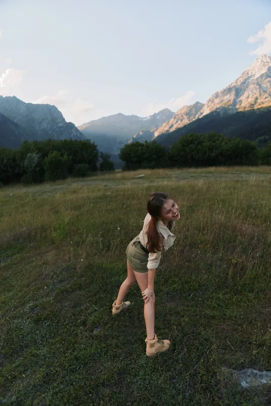 Young woman in a light hiking outfit stands in a wide grassy field with towering mountains in the background, enjoying a carefree outdoor moment.