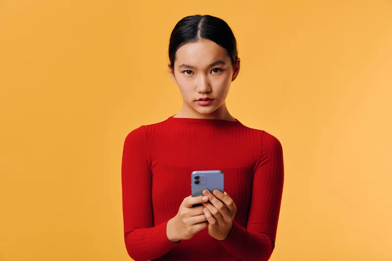 Young woman in red sweater looking at camera holding smartphone in both hands on orange background. Portrait of focused Asian female using mobile device for communication or technology.