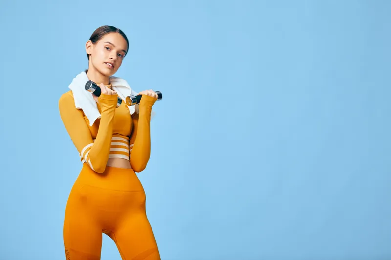 Athletic woman in bright orange workout set with towel draped over shoulders, holding a dumbbell across her shoulders against a blue studio backdrop, showcasing strength, fitness and sport.