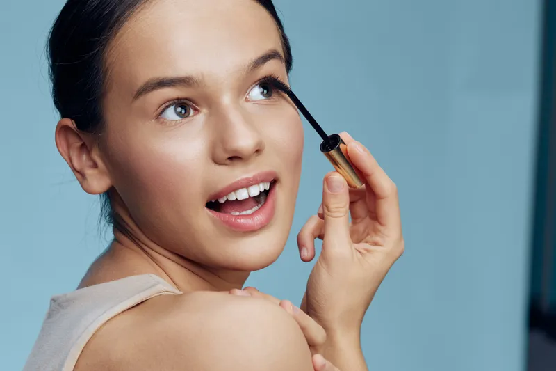 Young woman applying mascara with a joyful expression, showcasing beauty and confidence against a soft blue background in a skincare concept