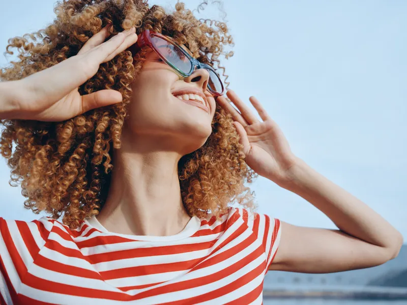 Portrait of a joyful woman with curly hair, wearing a red and white striped shirt and sunglasses, basking in bright sunlight against a clear blue sky.