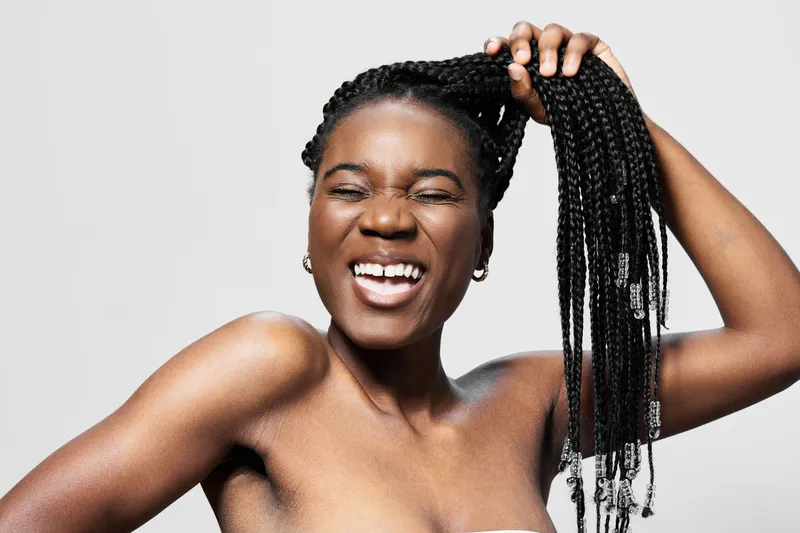 Happy young woman with braids playfully pulling her hair, joy and confidence expressed against a minimalist grey background