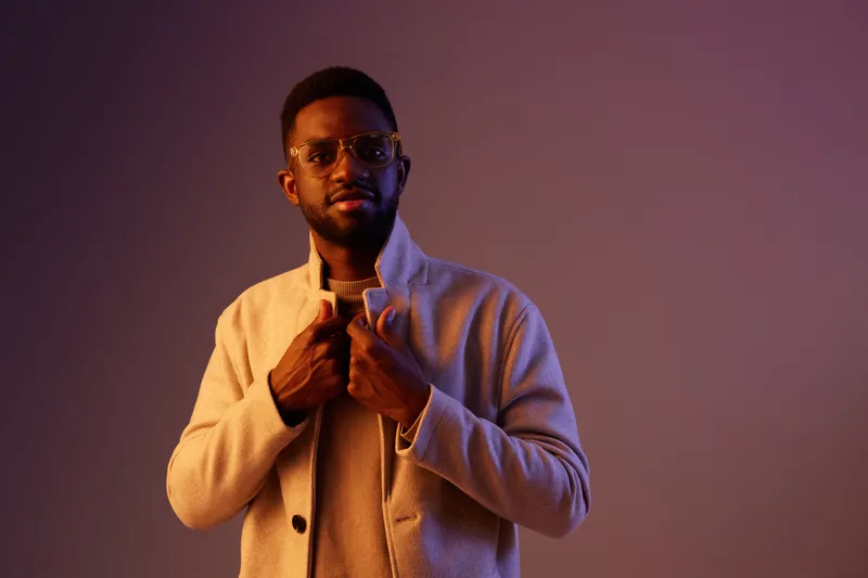 Confident young Black man in stylish outfit posing against a gradient purple background, exuding a modern and fashionable vibe