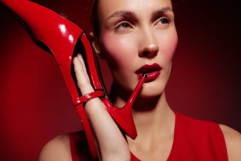 Close-up of a woman holding a red high heel shoe near her face against a red background. Concept of fashion, style, glamour, and bold makeup with red lipstick and blush.