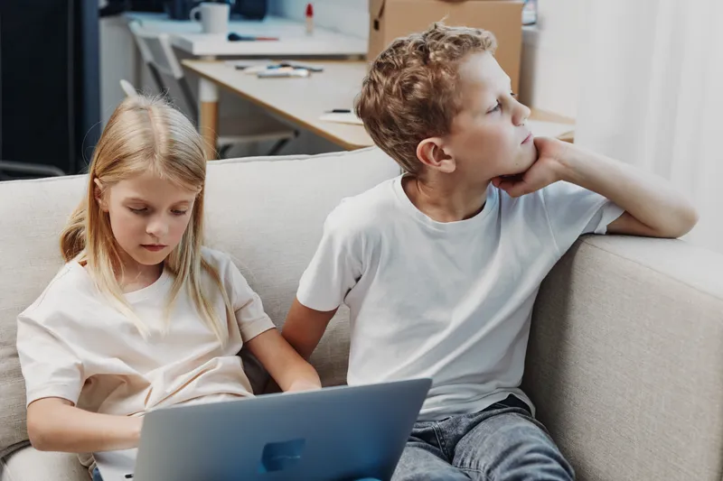 Two children sit on a couch, a girl using a laptop focused while the boy looks away thoughtfully in a modern home interior.