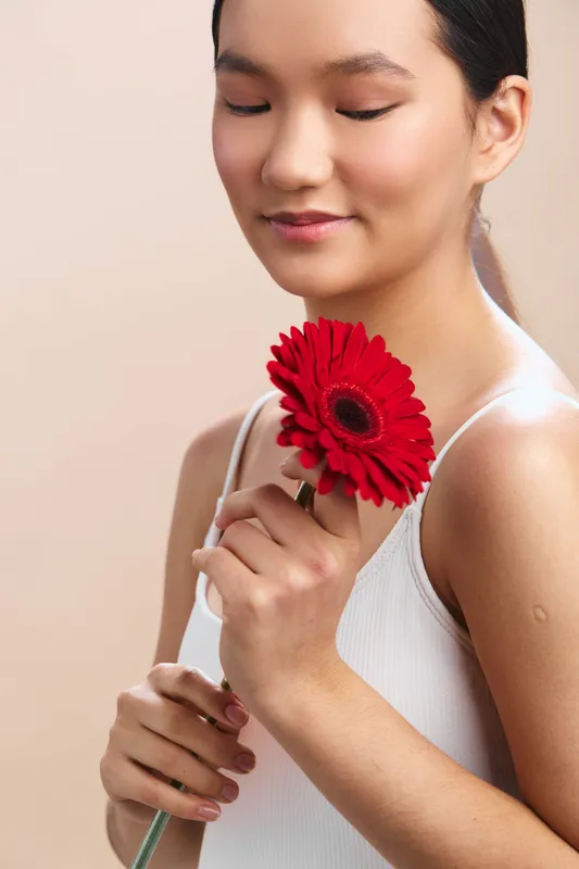asian woman in a sleeveless white top holds a vibrant red flower near her face, her eyes closed with a gentle smile, expressing calm grace and quiet joy in a soft studio setting.