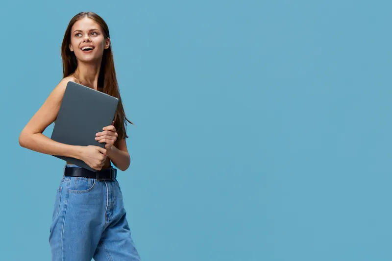 smiling woman holding laptop confidently wearing casual blue jeans standing against plain blue background with natural light and cheerful expression