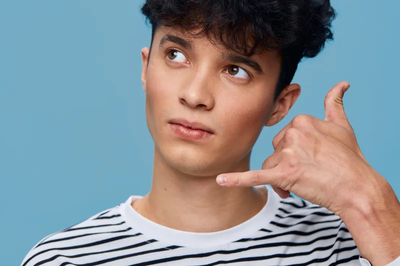 Young man with curly hair wearing striped shirt making call me gesture against blue background, thoughtful expression and casual style in studio lighting.