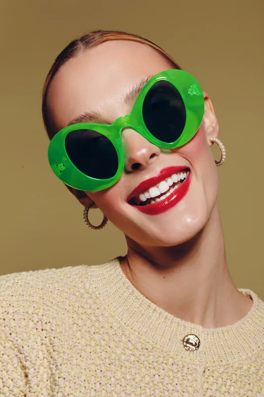 Smiling woman with bright red lipstick wears large green sunglasses and gold hoop earrings. She poses in a beige textured sweater against a neutral background. Fashion and style concept.