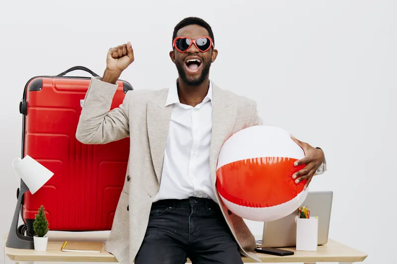 Happy young man with dark hair and beard wearing sunglasses, white shirt, and beige blazer holding a beach ball, standing next to a large red suitcase in a bright studio setting. People lifestyle vaca