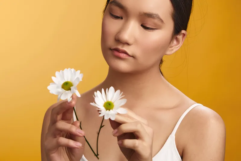 Asian woman gently holds white daisies against a warm yellow background, focusing on delicate petals and a serene expression to convey natural beauty and calm mood.