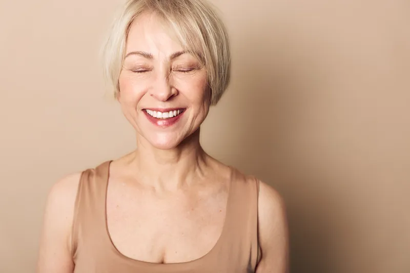 Smiling mature woman with closed eyes and short blonde hair wearing beige top stands against plain beige background. Concept of happiness, serenity, confidence, natural beauty, and peaceful moments.