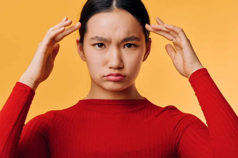 Young woman with long dark hair in red sweater shows frustrated expression holding temples, isolated on orange background with serious mood and focused look.