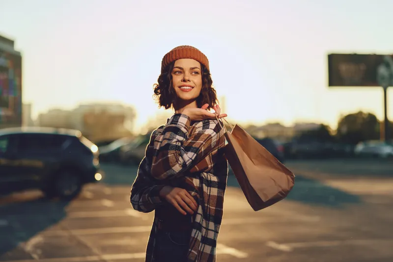Woman shopping with smile at sunset in urban parking, wearing beanie and plaid coat holding a bag in candid moment; authenticity, mindful living and golden hour glow with emotional storytelling.