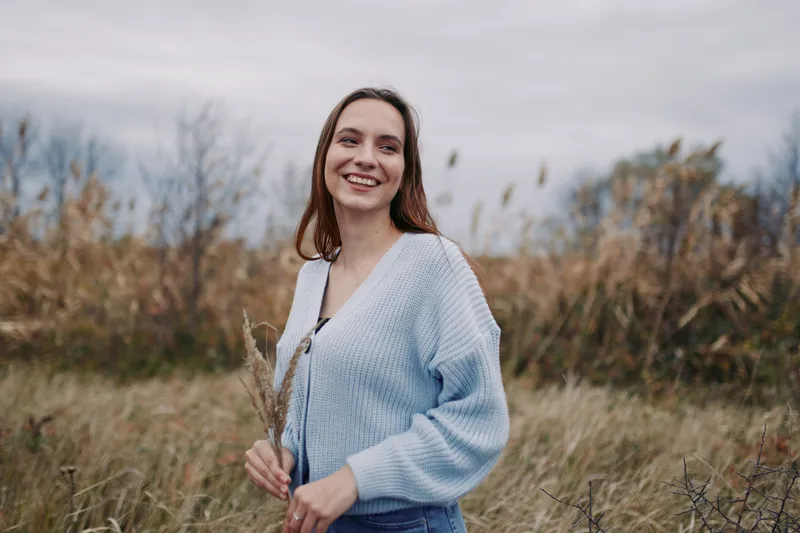 Young woman smiles in a windswept meadow holding grasses in a cozy sweater; her genuine expression and relaxed posture convey authenticity and credibility in a candid portrait.