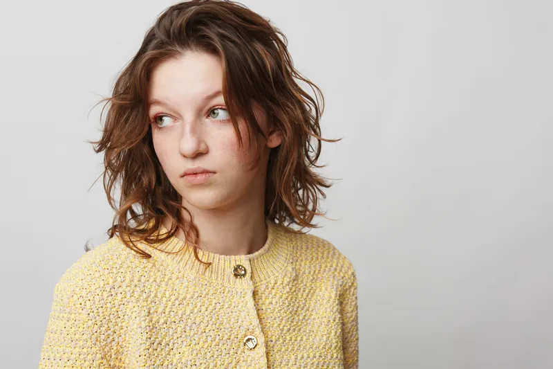 Young woman with curly hair wearing yellow knitted sweater looks away thoughtfully on grey background. Concept of contemplation, mood, casual style, and natural beauty portrait.