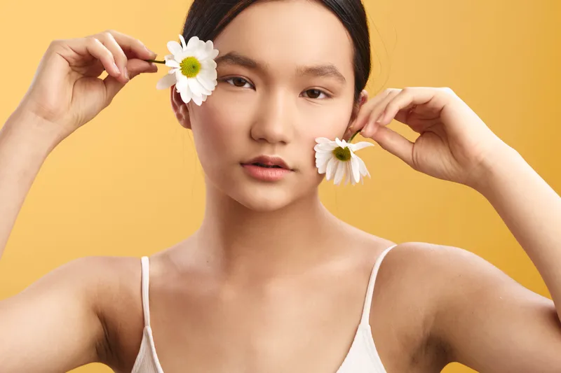 Asian woman with flowers frames her face in a bright studio, exuding calm confidence and natural beauty against a vibrant backdrop. Her serene expression with soft lighting creates a contemporary