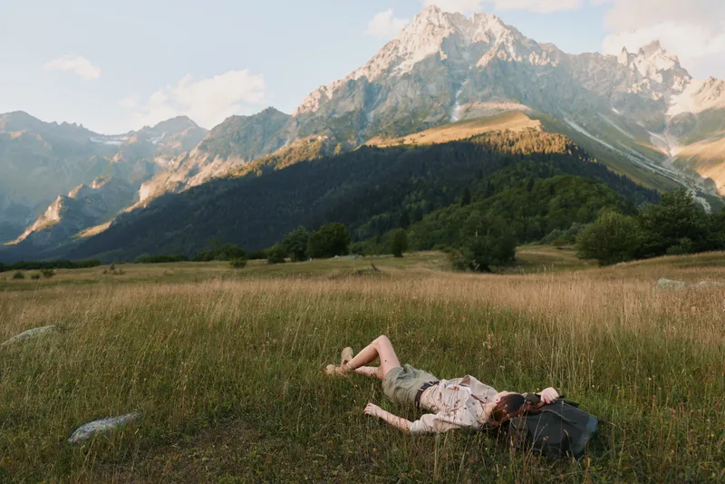 Young woman lies on a grassy field in casual clothes resting with a backpack near her head against a backdrop of sunlit mountains under a partly cloudy sky.