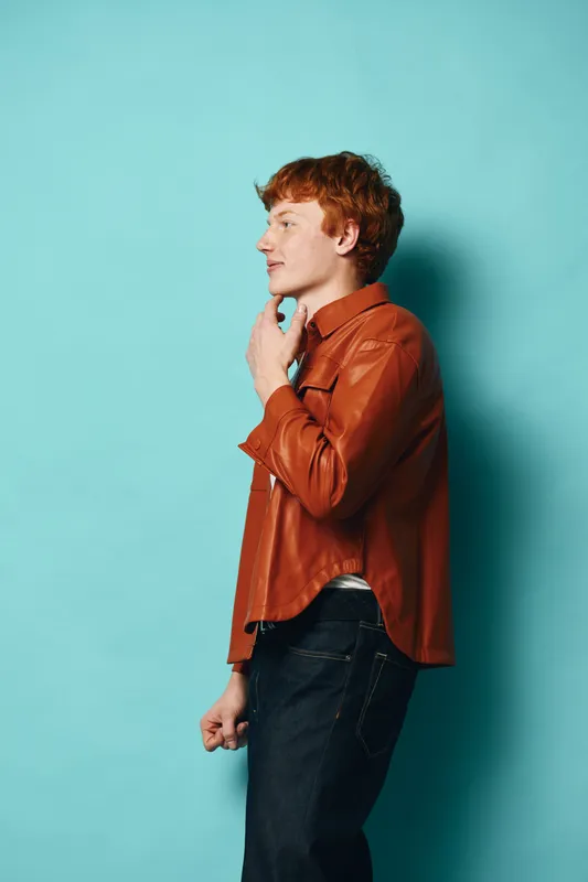 Young man with red hair wearing brown leather jacket and jeans standing in profile against blue background touching chin in thoughtful pose for casual style portrait.