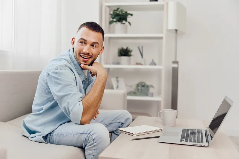Smiling man casually dressed in a light blue shirt, seated in a modern living room, engaged in a productive work session with a laptop and notebook.