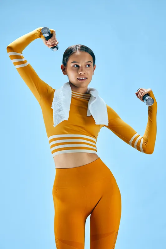 Female fitness enthusiast in bright orange athletic wear lifts dumbbells with a towel around her neck, dynamic pose against a blue backdrop, conveying energy, focus and empowered workout mindset.