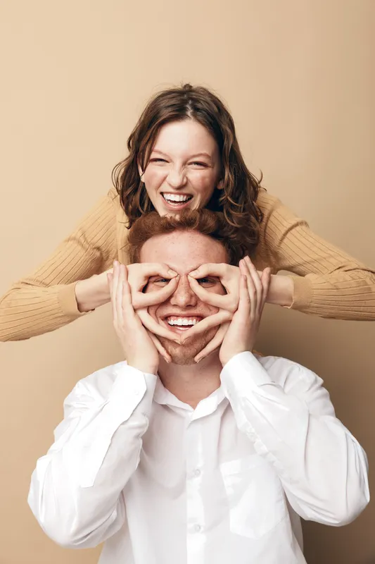 Happy young couple having fun together, woman playfully covering man's eyes with hands. Studio portrait on beige background. Concept of love, joy, friendship, and positive emotions.