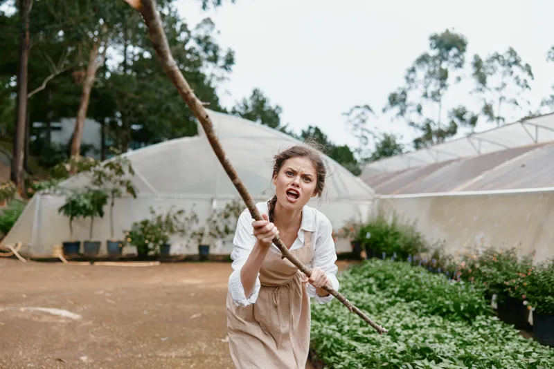 Young woman in a greenhouse, expressing frustration while holding a stick, dressed in casual attire with a natural background