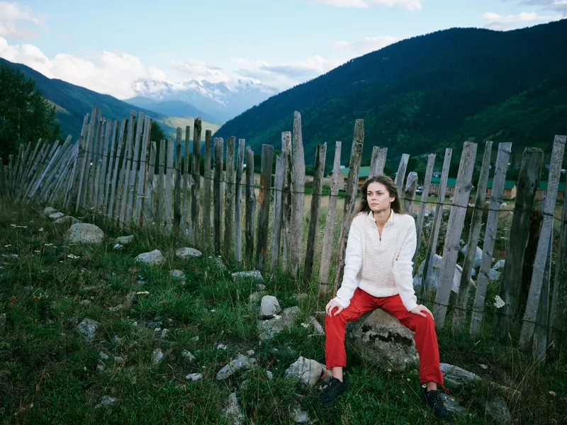Woman sits in the countryside beside a weathered fence, mountains in the distance and open scenery around a rocky valley, wearing a casual white top and red pants amid a green landscape and tranquil