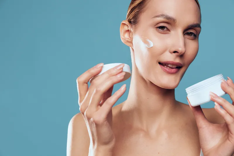 Young woman applying face cream with fingers, skincare routine, clear skin, natural beauty, soft lighting, blue background, moisturizing product, beauty care concept