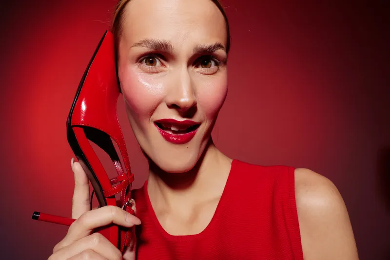 Close-up of a smiling woman in red dress holding a red high heel shoe near her face on red background. Concept of fashion, style, glamour, and confident beauty.