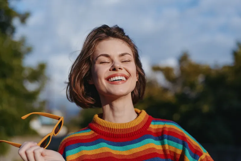 Woman smile portrait in colorful striped sweater holding sunglasses outdoors with eyes closed, enjoying warm sunlight and carefree happiness in a candid lifestyle headshot, young female model.