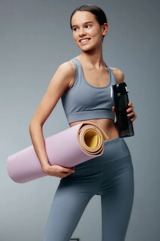 Fit woman in athletic wear holds a yoga mat and water bottle, ready for a workout. Sporty pose emphasizes fitness lifestyle, training mindset and healthy routine in a studio.