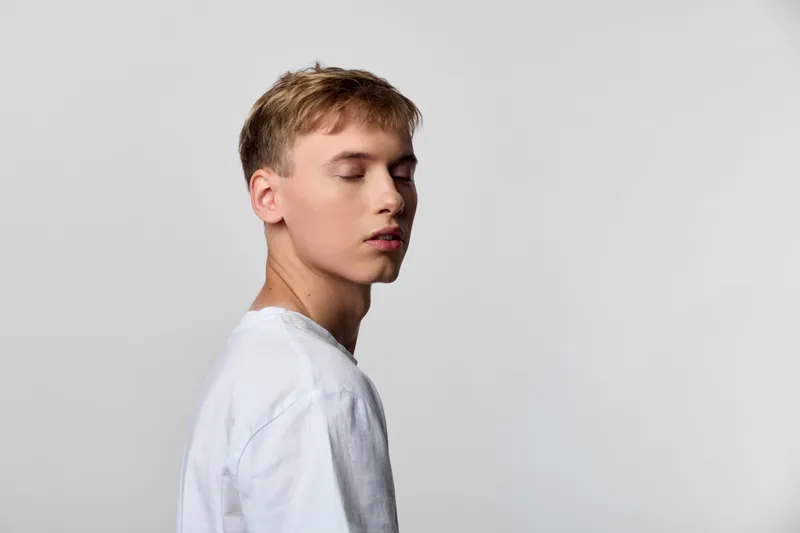 Young man in white shirt with closed eyes and serene expression, side profile against a plain light background, conveying calm, contemplation, and minimalist fashion mood for editorial use and