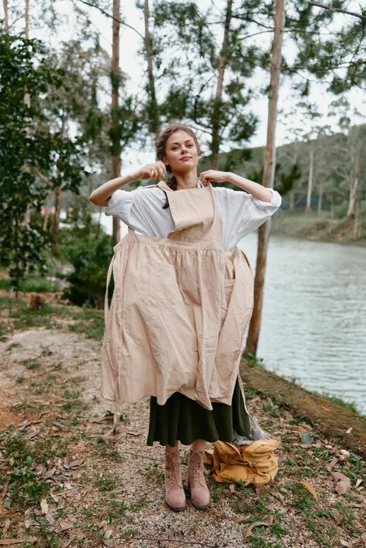 Young woman in nature wearing a beige apron preparing for gardening, showcasing a serene expression by the lakeside, with a soft color palette of greens and browns