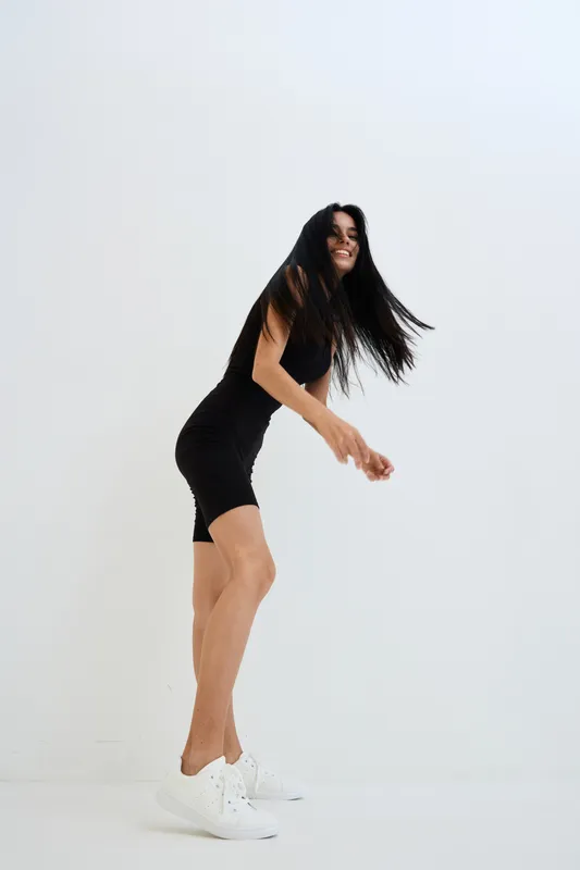 Woman in black fitted outfit jumping joyfully with long hair flowing, white sneakers, and a clean minimalist white background, expressing energy and happiness.